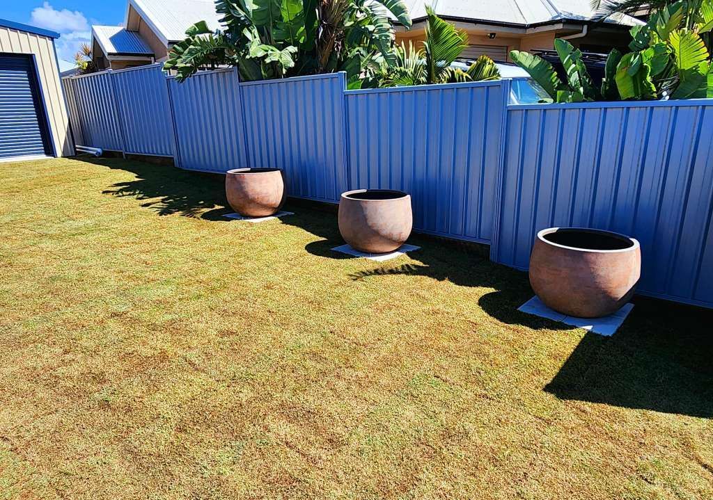 Three Brown Planters on Gray Squares in a Grassy Yard Next to a Blue Fence — On The Mark Landscaping in Branyan, QLD
