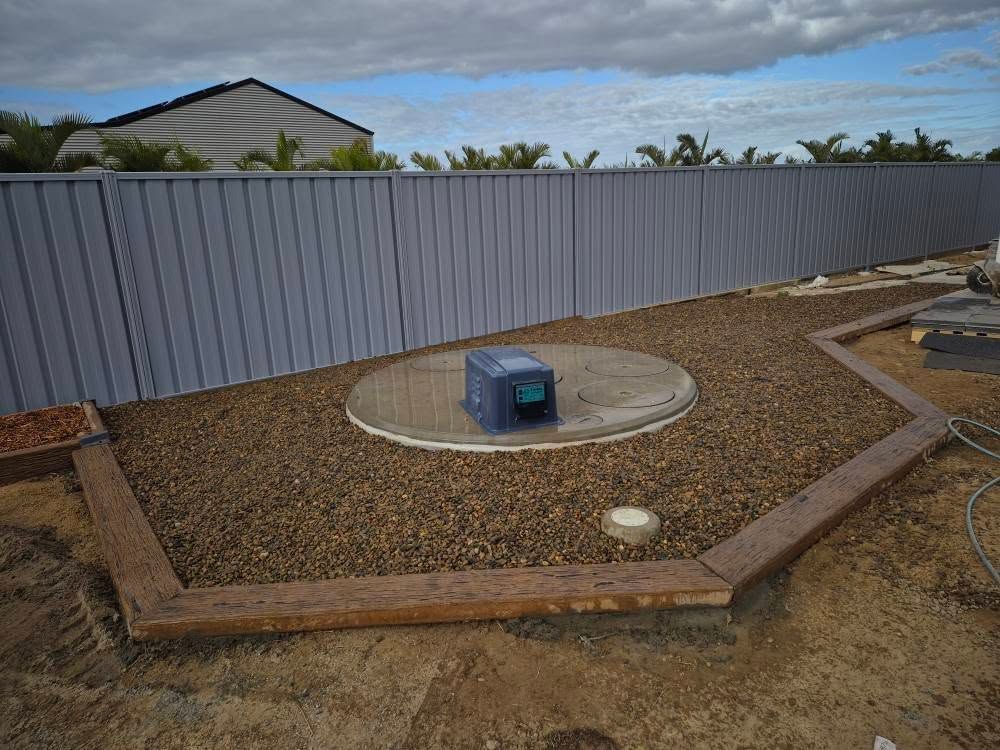 A Gravel Bed With a Concrete Circle, Machinery, Wooden Planks and a Grey Fence — On The Mark Landscaping in Branyan, QLD