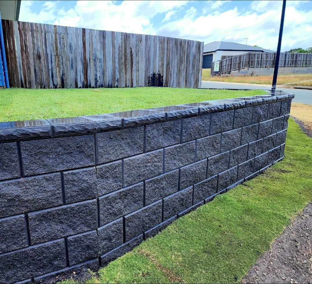 Gray brick retaining wall with green grass and wooden fence in the background — On The Mark Landscaping in Branyan, QLD