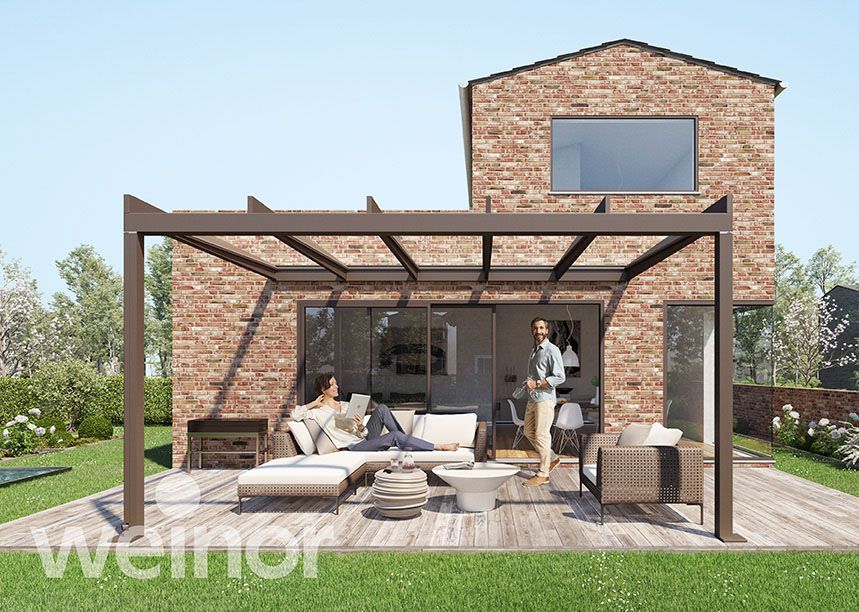 Eine Frau sitzt auf einem Sofa unter einer Pergola vor einem Backsteinhaus.