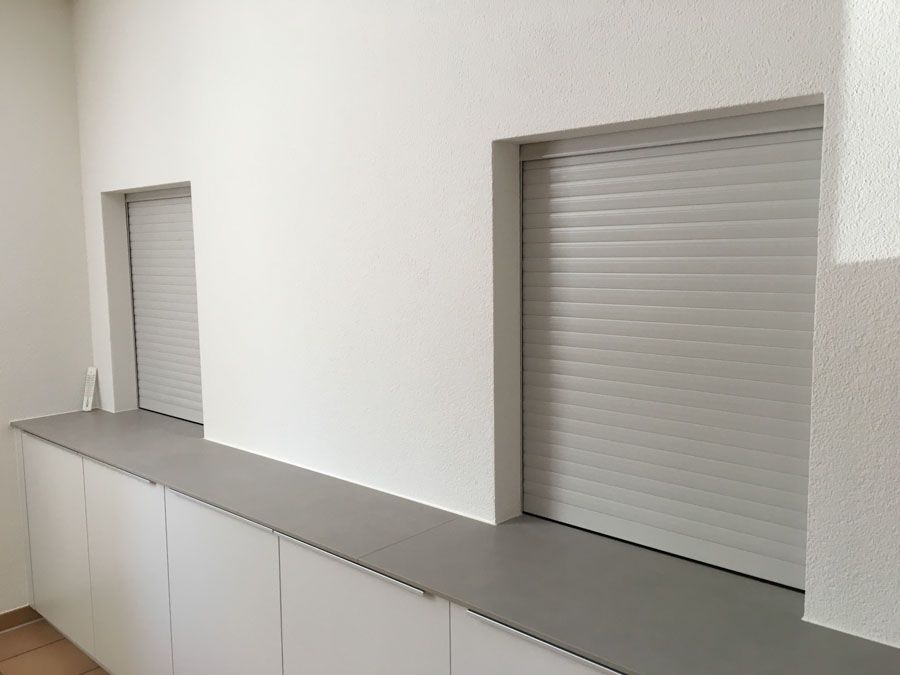 Ein Raum mit einer Reihe weisser Schränke und zwei Fenstern mit Rollläden.