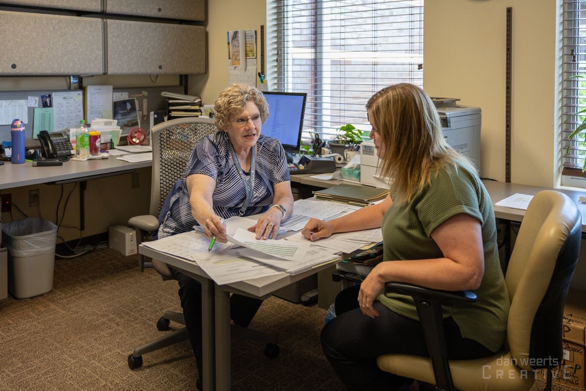 Two women are sitting at a table in an office talking to each other.
