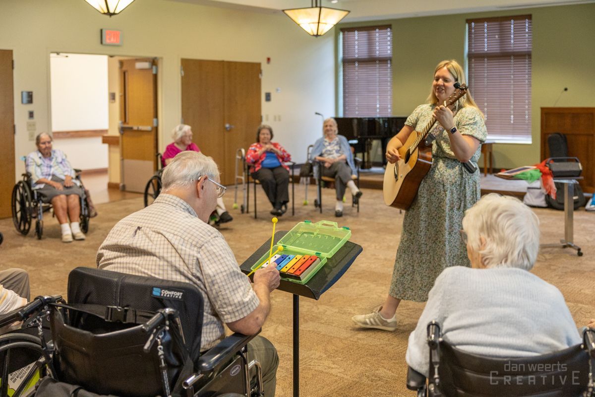 A nurse wearing a mask is talking to an elderly woman sitting on a bed.