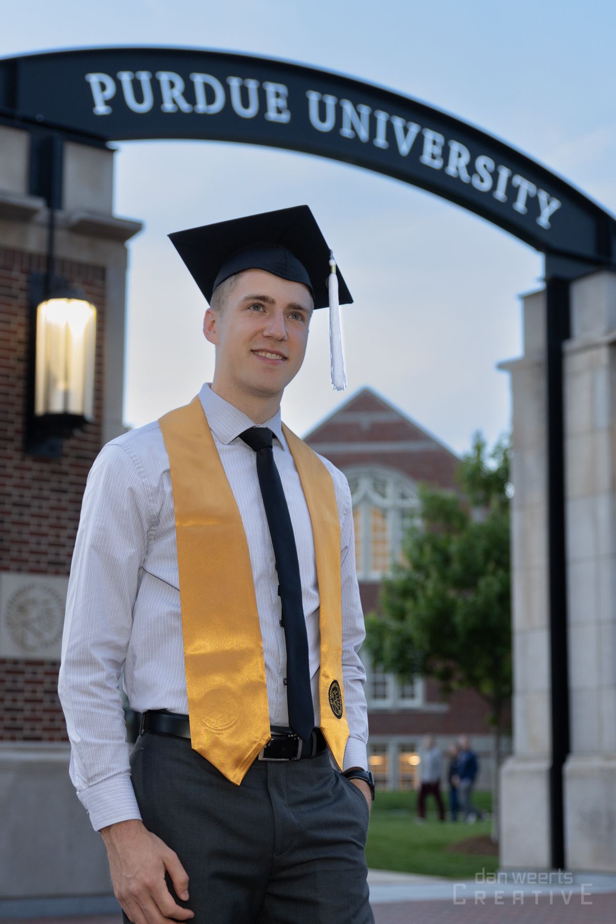 A man in a graduation cap and gown is standing in front of a purdue university archway.