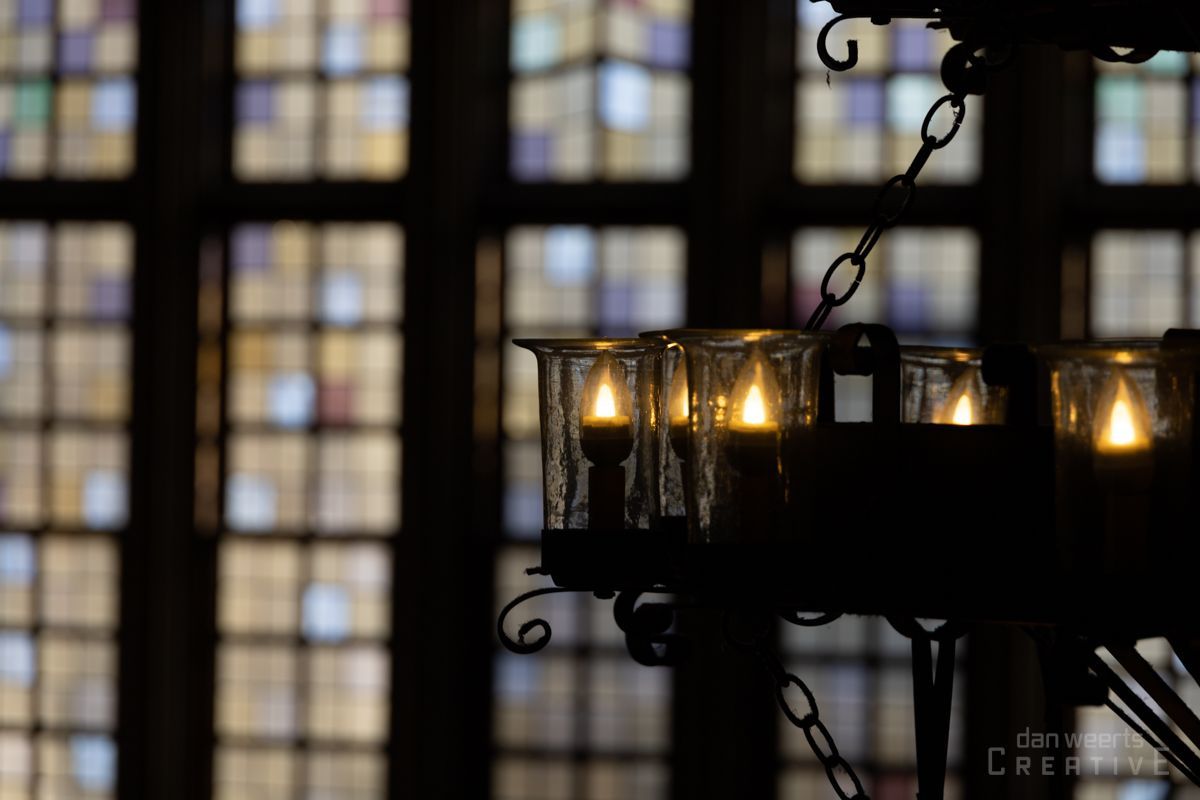 A chandelier with candles hanging from the ceiling in front of a stained glass window.