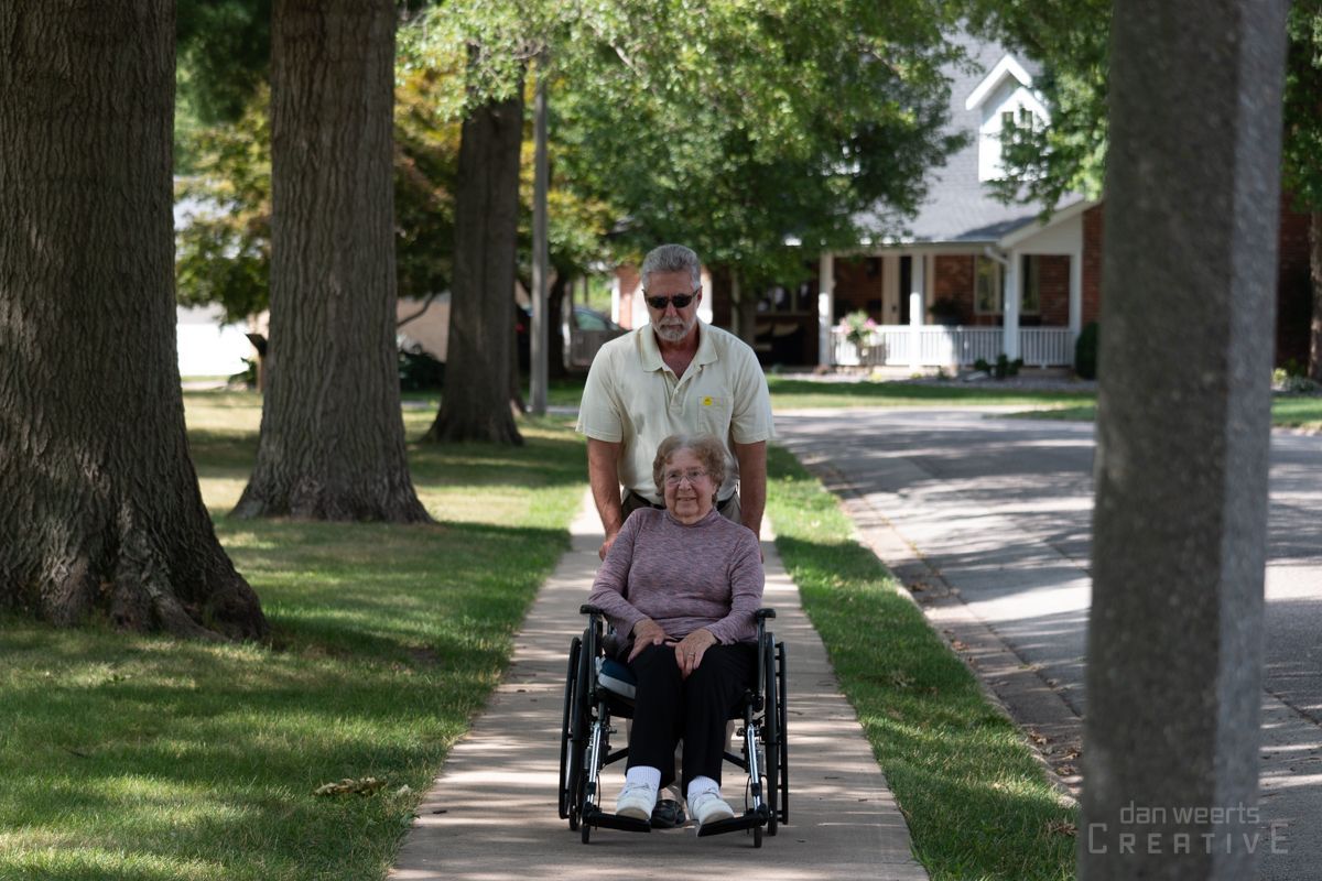 A man pushes a woman in a wheelchair down a sidewalk