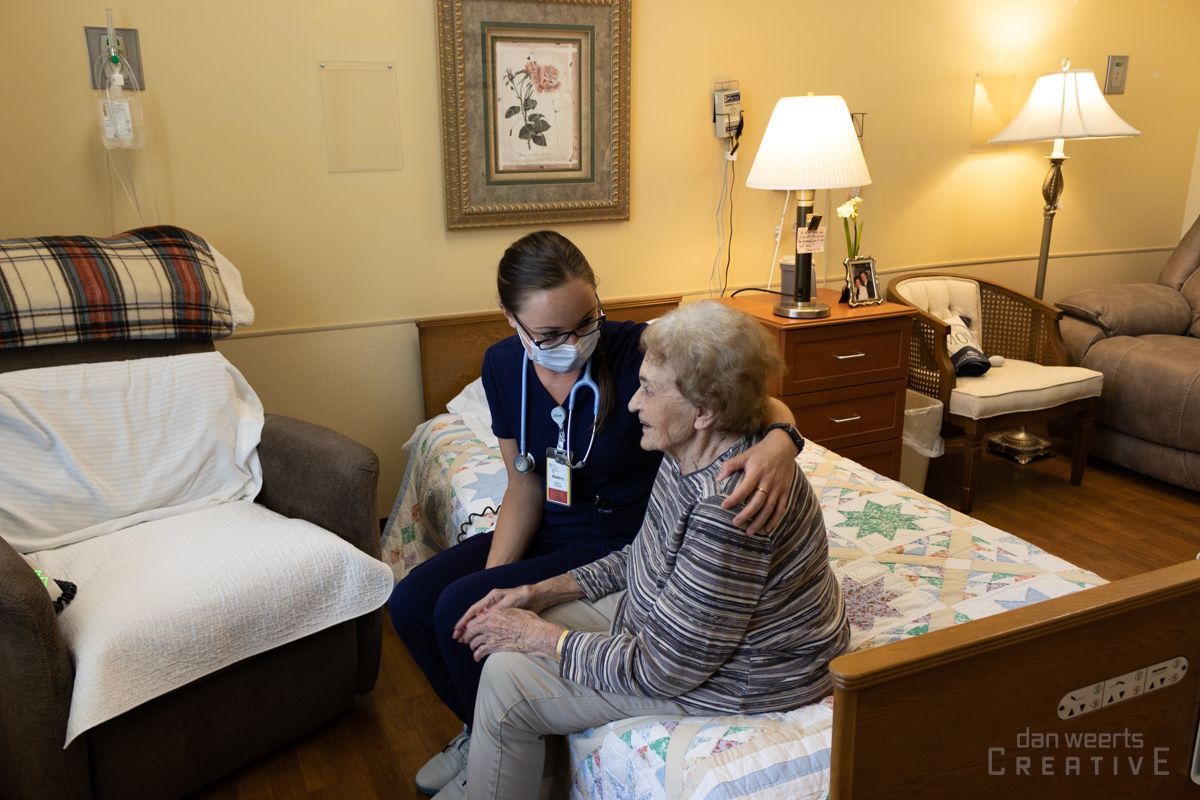 A nurse wearing a mask is talking to an elderly woman sitting on a bed.