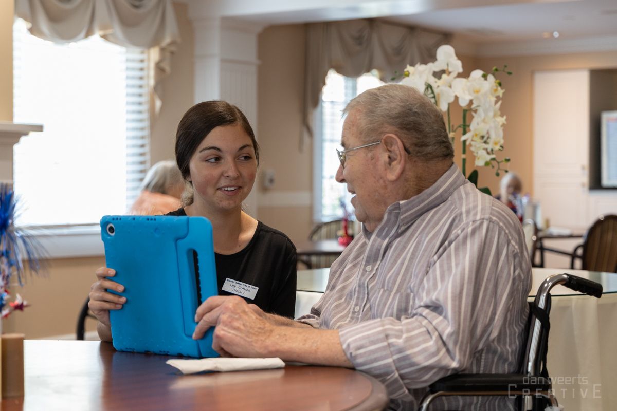 A woman is helping an elderly man in a wheelchair use a tablet computer.
