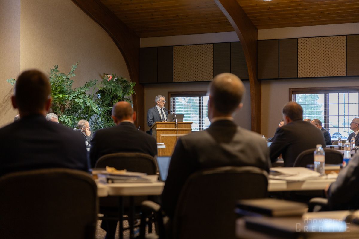 A group of people are sitting at tables in a room with a man standing at a podium giving a speech.