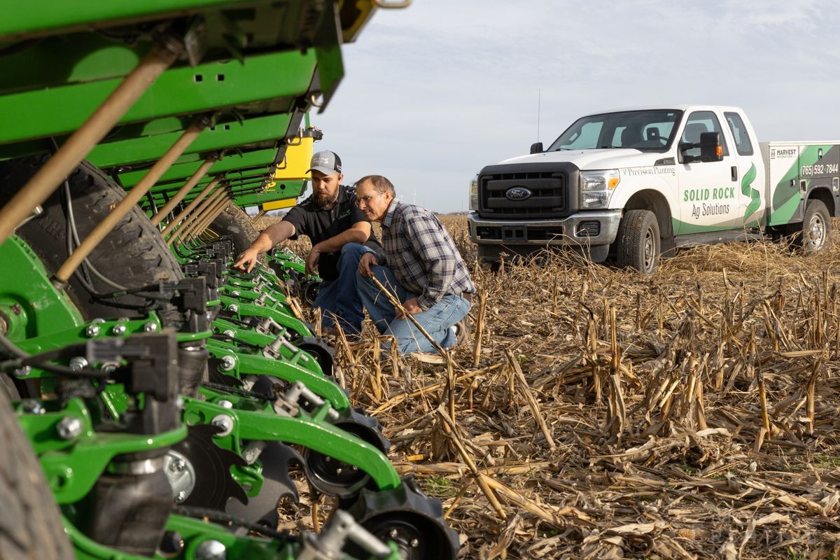 Two men are looking at a planter in a field with a truck in the background.