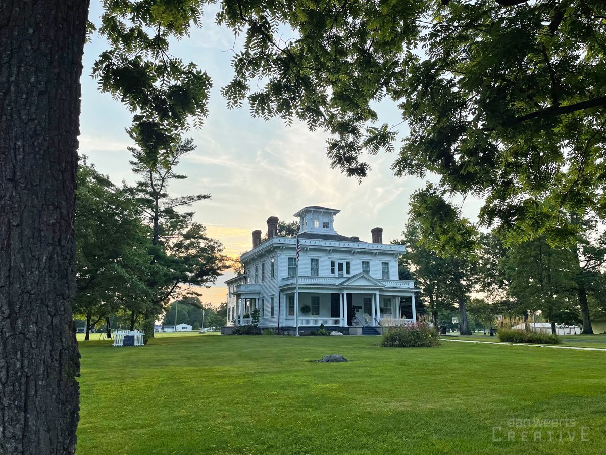 A large white house sits in the middle of a lush green field