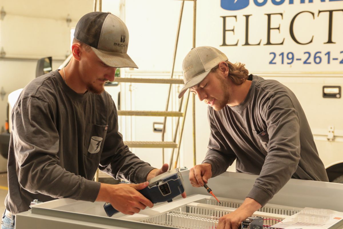 Two men are working on a piece of equipment in front of a sign that says elec