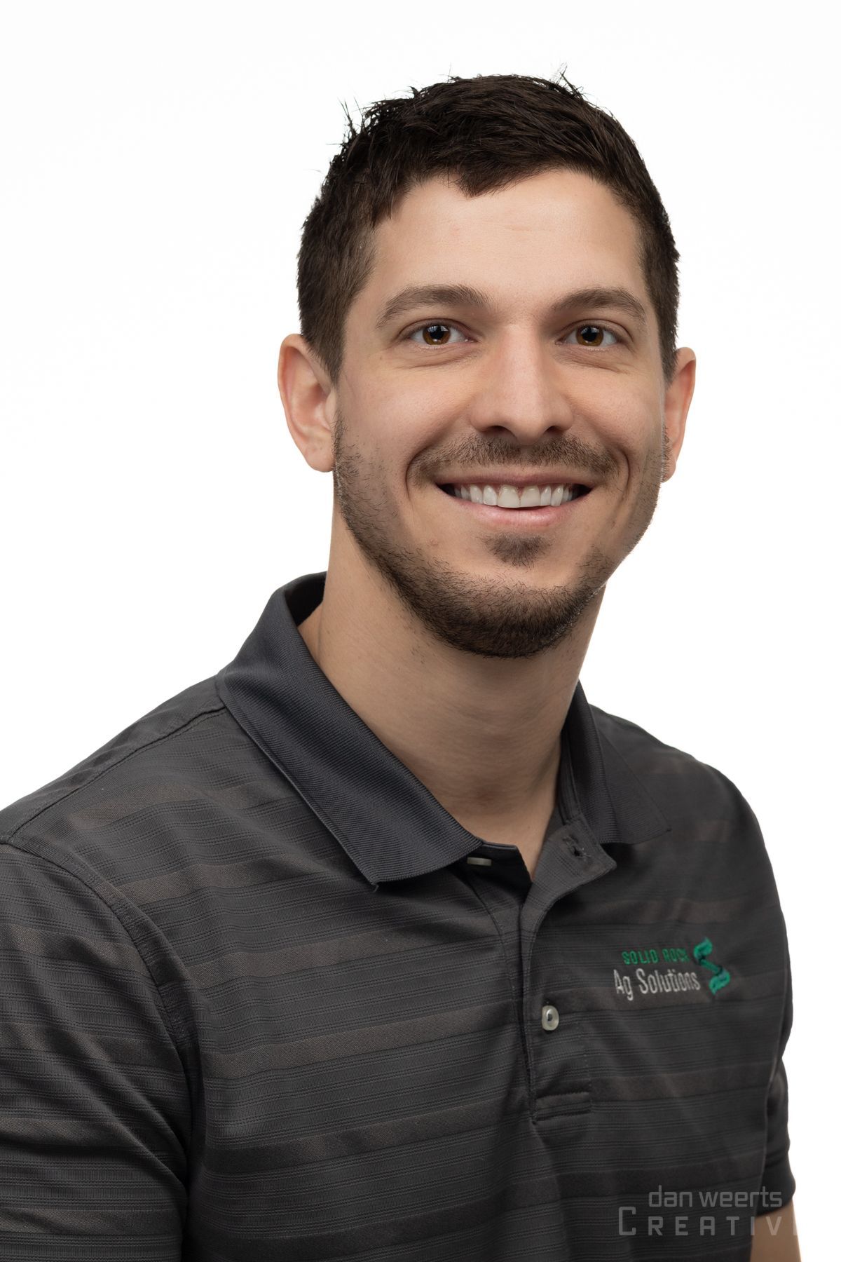 A man with a beard is smiling for the camera while wearing a black polo shirt.