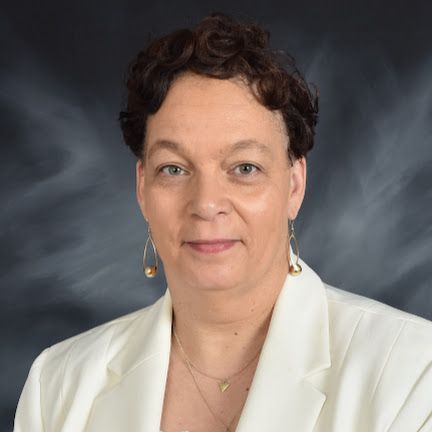 A woman wearing a white jacket and earrings is standing in front of a black background.