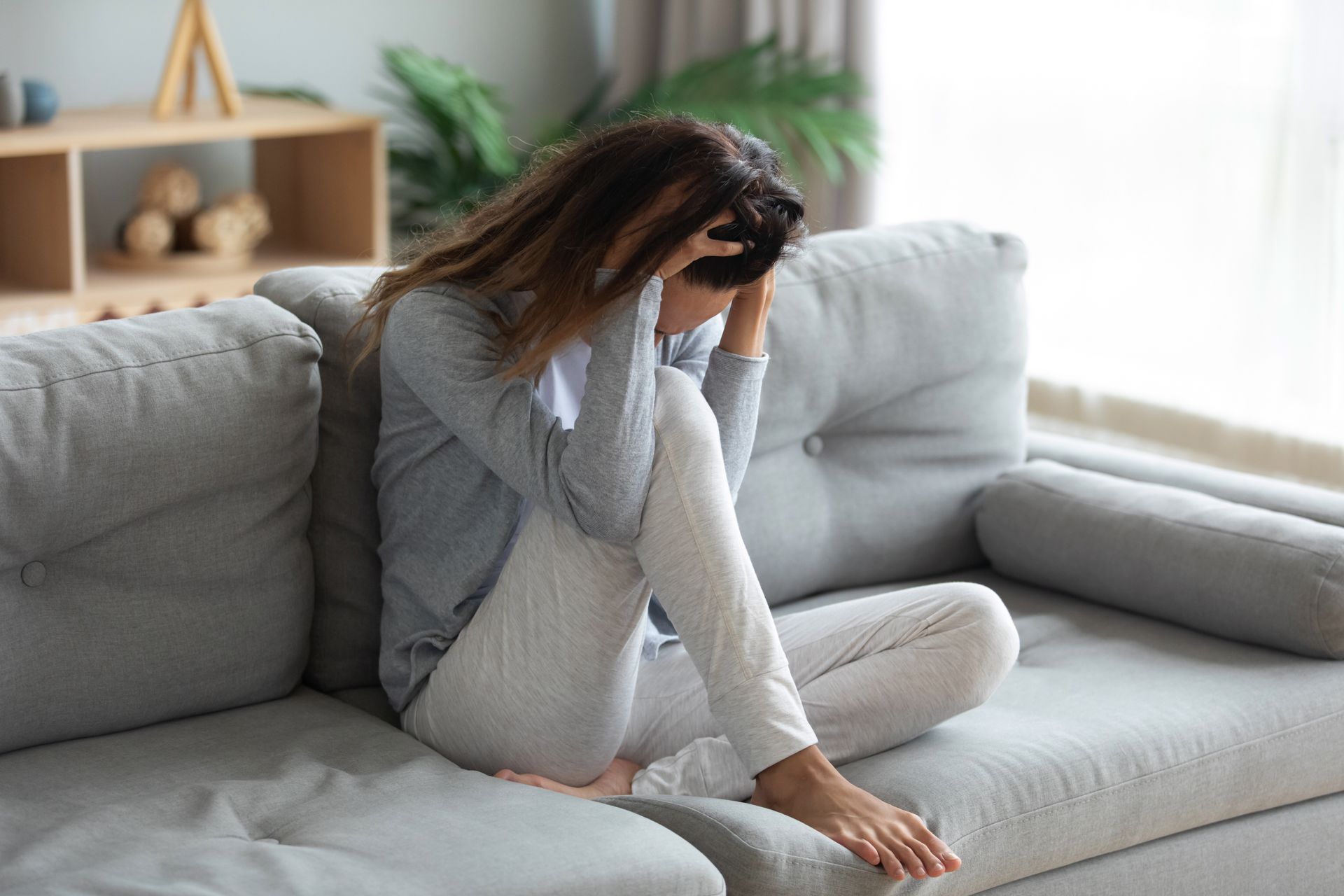 Woman sitting on a gray sofa, head in hands, looking upset in a bright living room