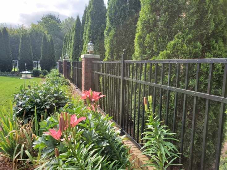 Black metal fence with brick pillars, lined with evergreen trees and flower garden.