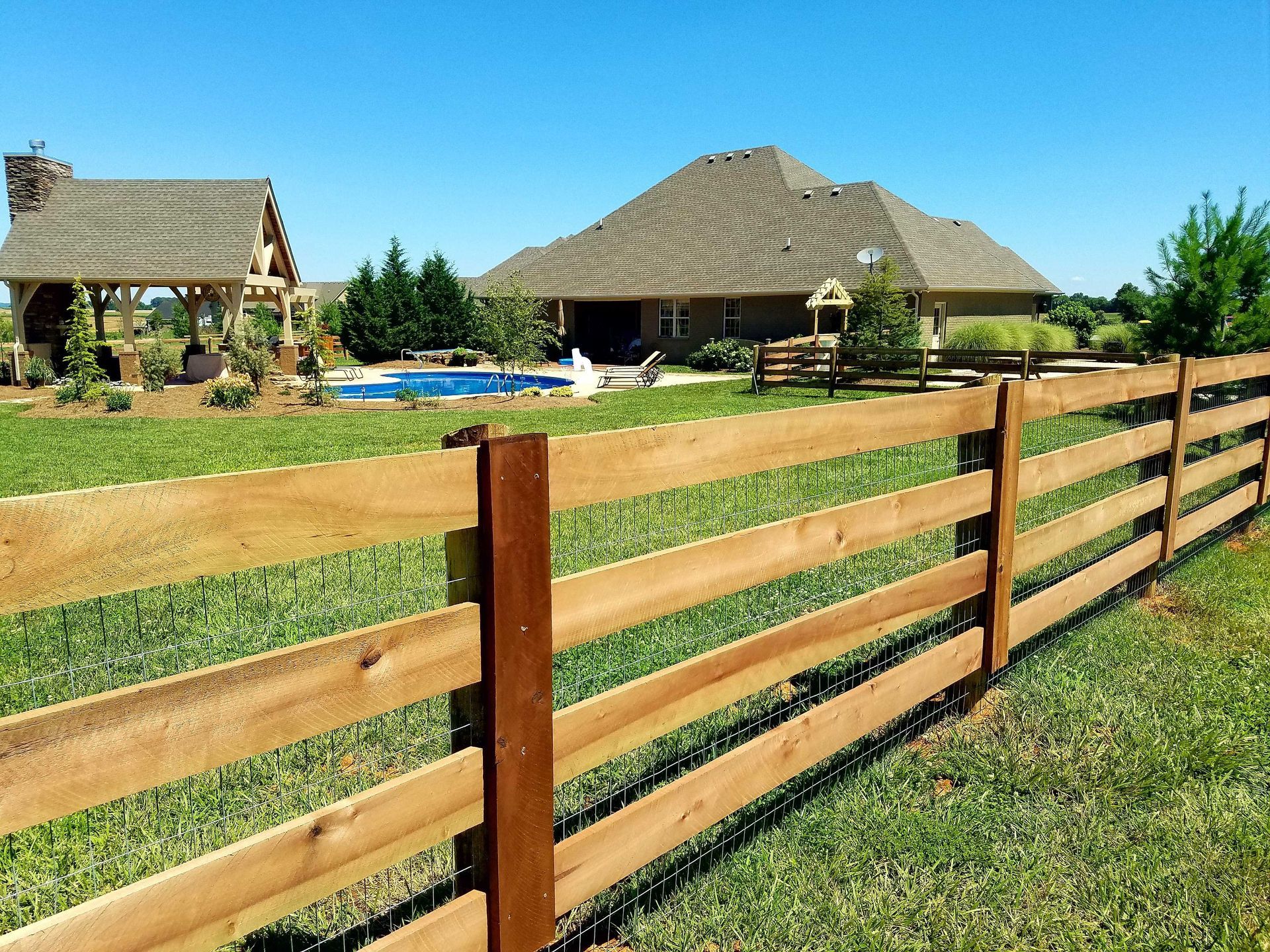Wooden fence in front of a green lawn with a house, a pool, and a gazebo on a sunny day.