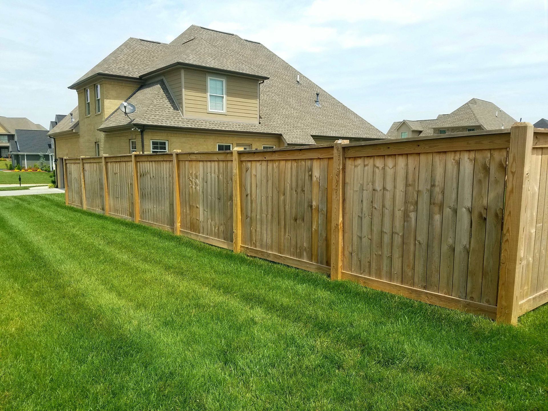 Wooden fence bordering a lush green lawn next to a two-story beige house under a blue sky.