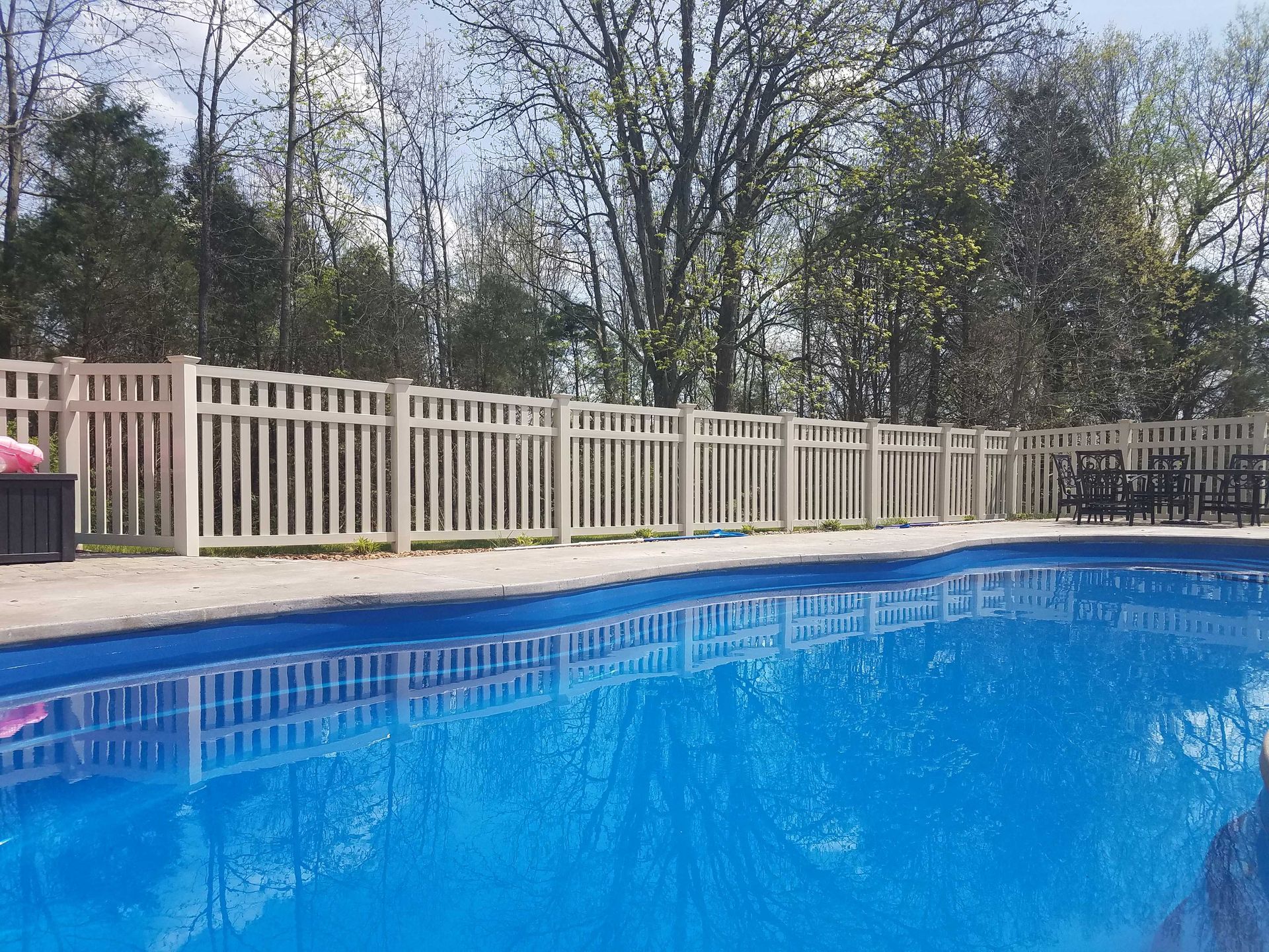 Blue swimming pool with a cream-colored fence in a backyard, trees in the background.