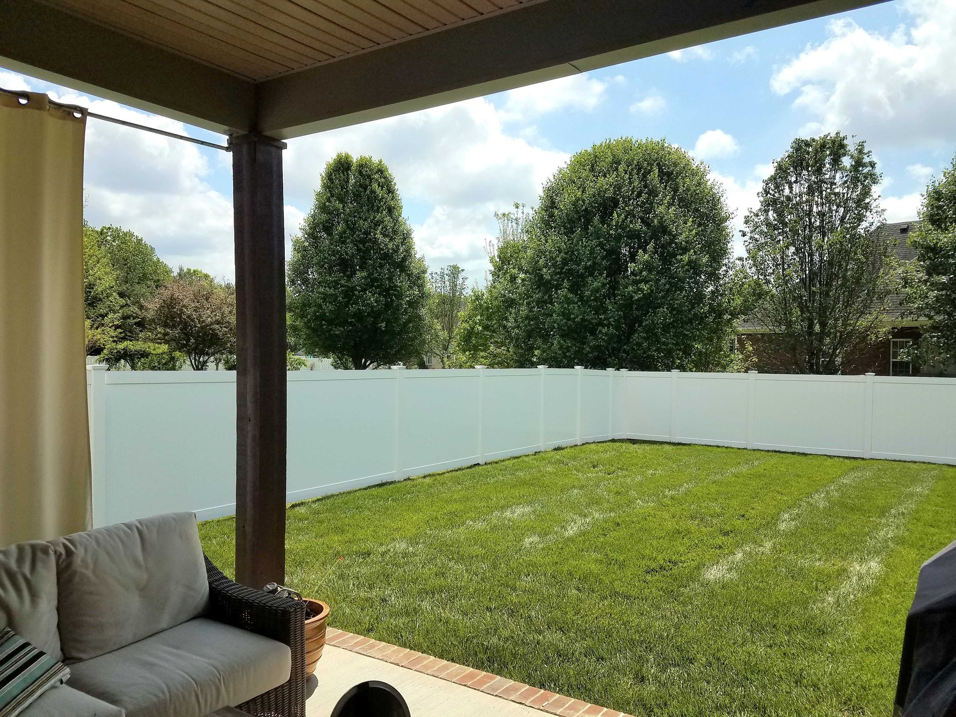 A backyard with a white fence, green grass, trees, and a covered patio with a couch.