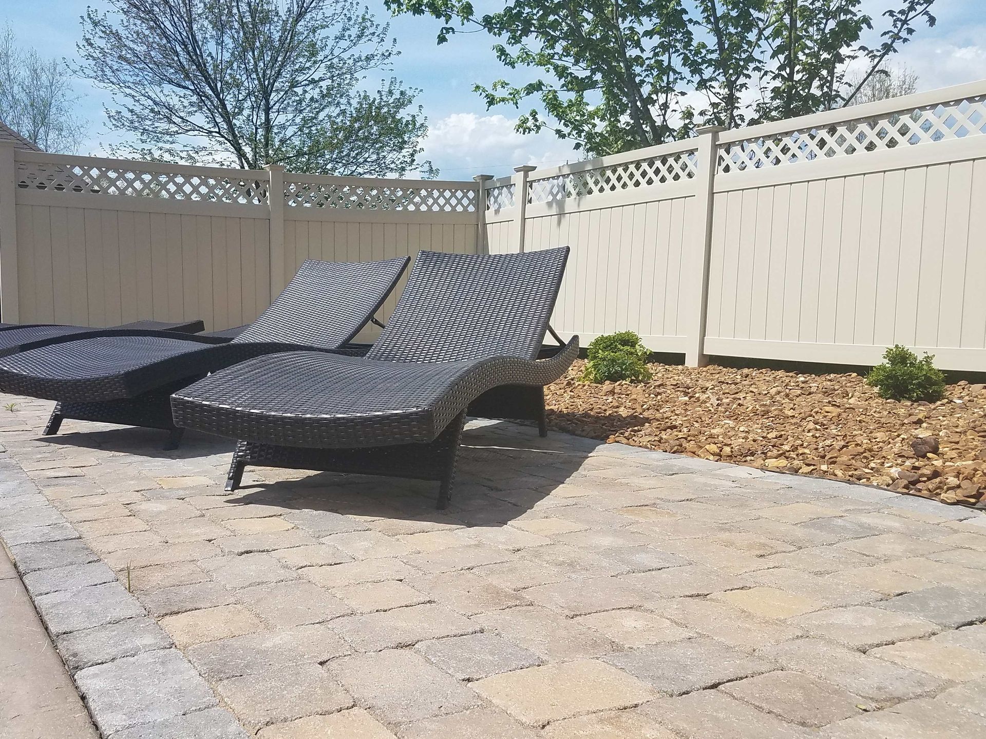 Lounge chairs on stone patio by a lattice fence. Pebbles and greenery in background.