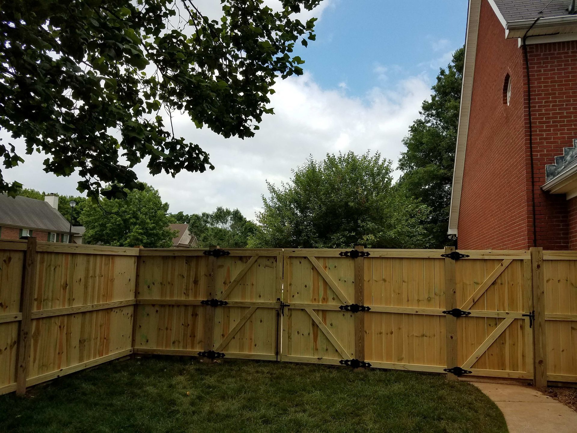 Wooden fence encloses a green lawn, next to a red brick building, under a cloudy sky.