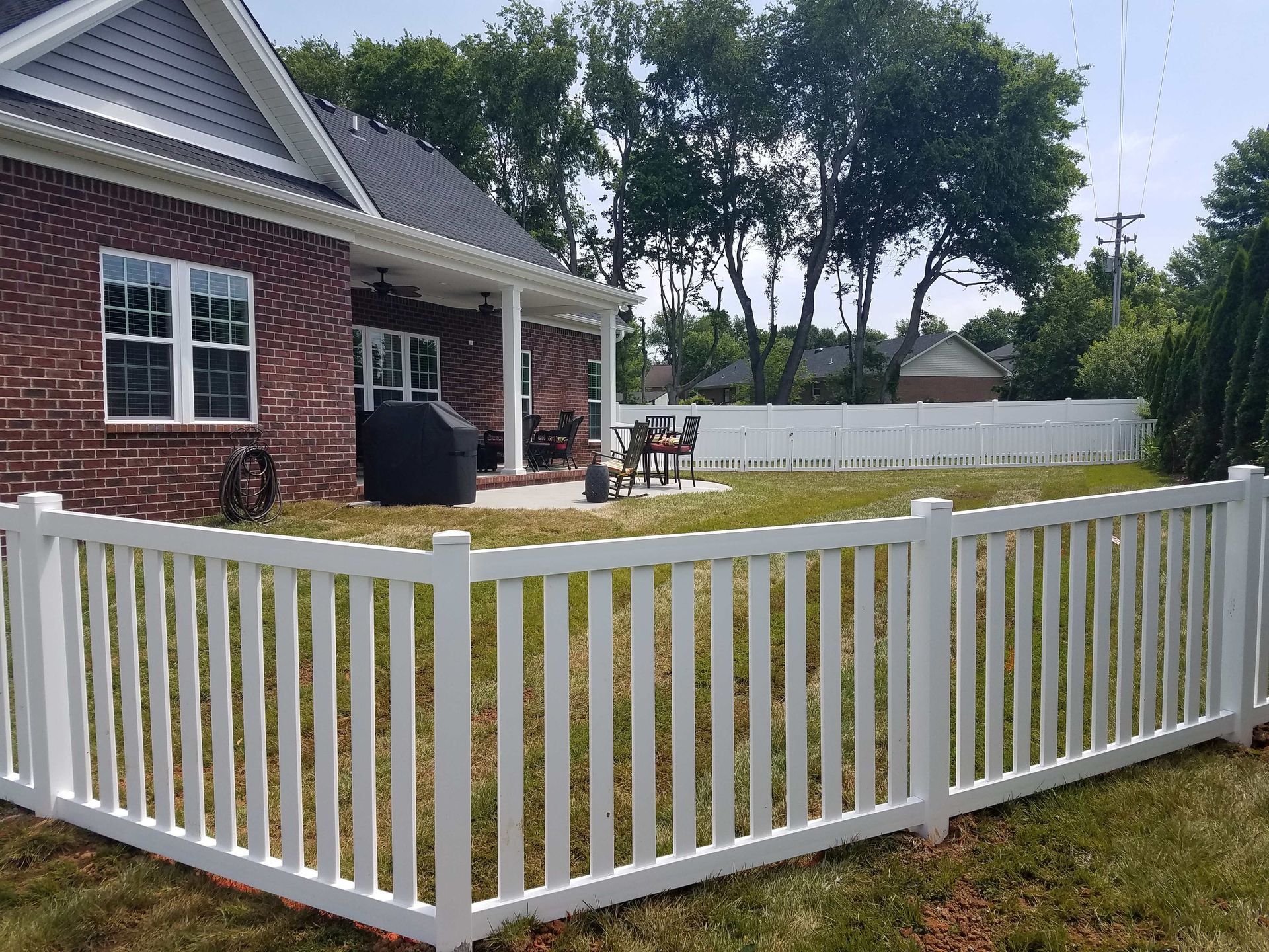 White picket fence encloses a green lawn with a red brick house in the background.