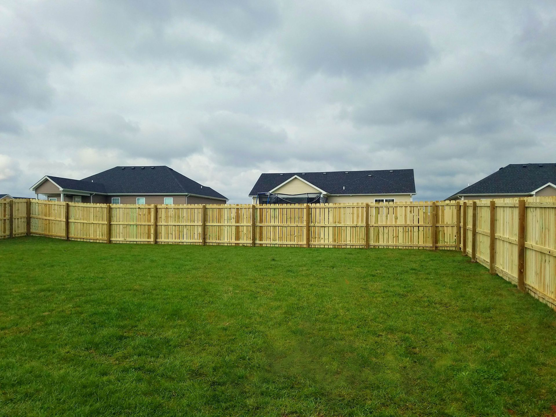 A grassy backyard with a wooden fence and houses in the background under a cloudy sky.