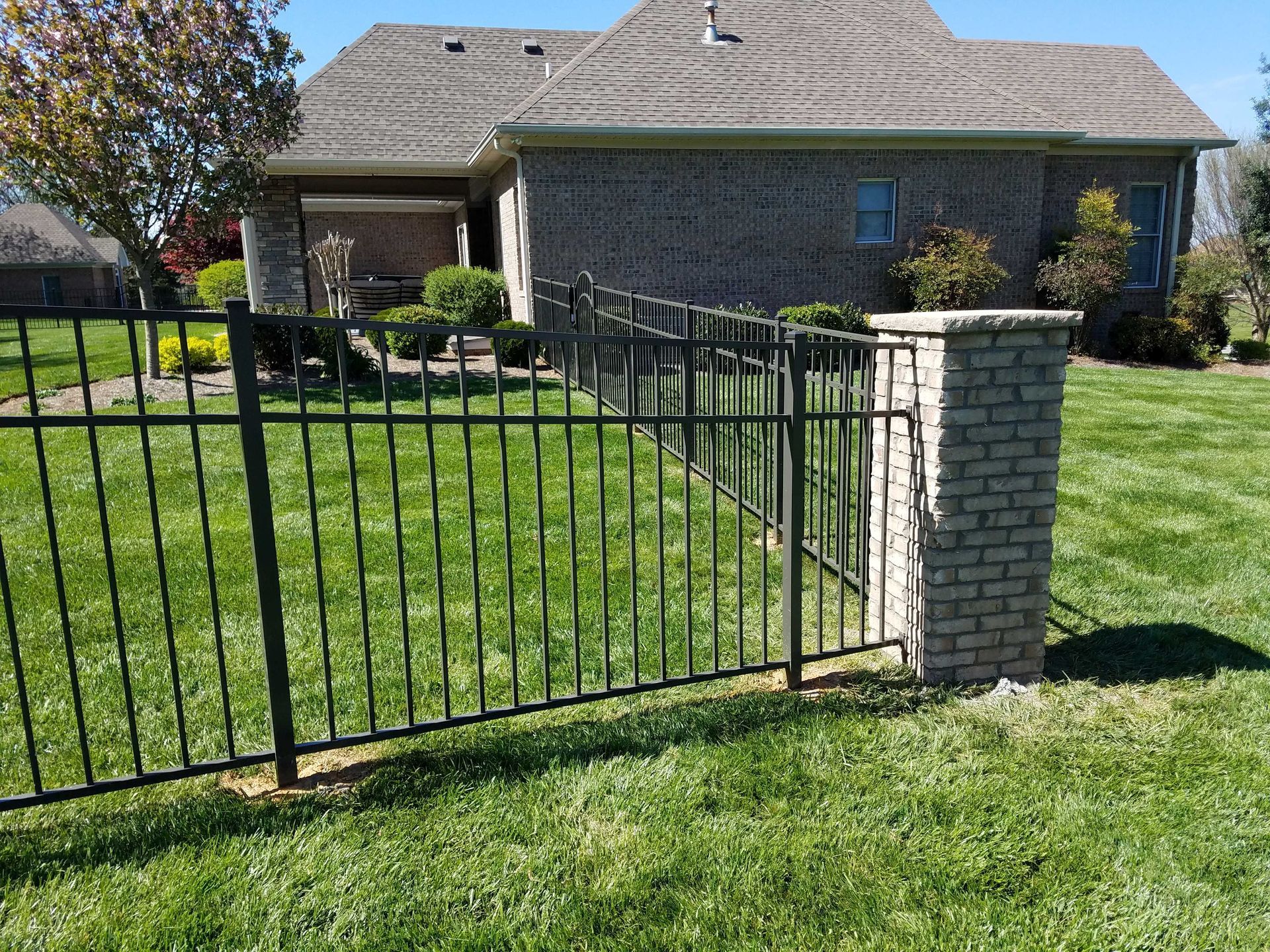 Black metal fence in a grassy yard, partially framing a brick house with a brown roof.