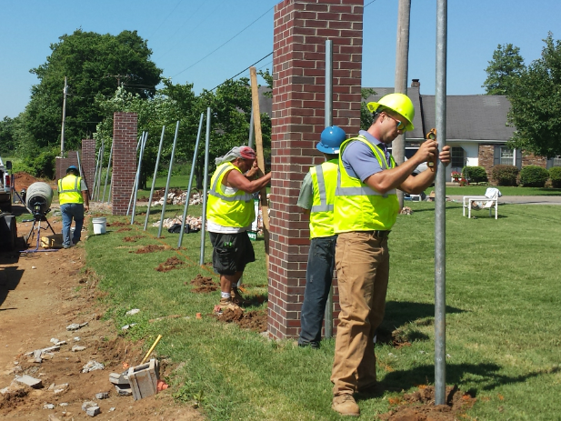 Construction workers installing metal fence posts near brick columns on a grassy lawn.