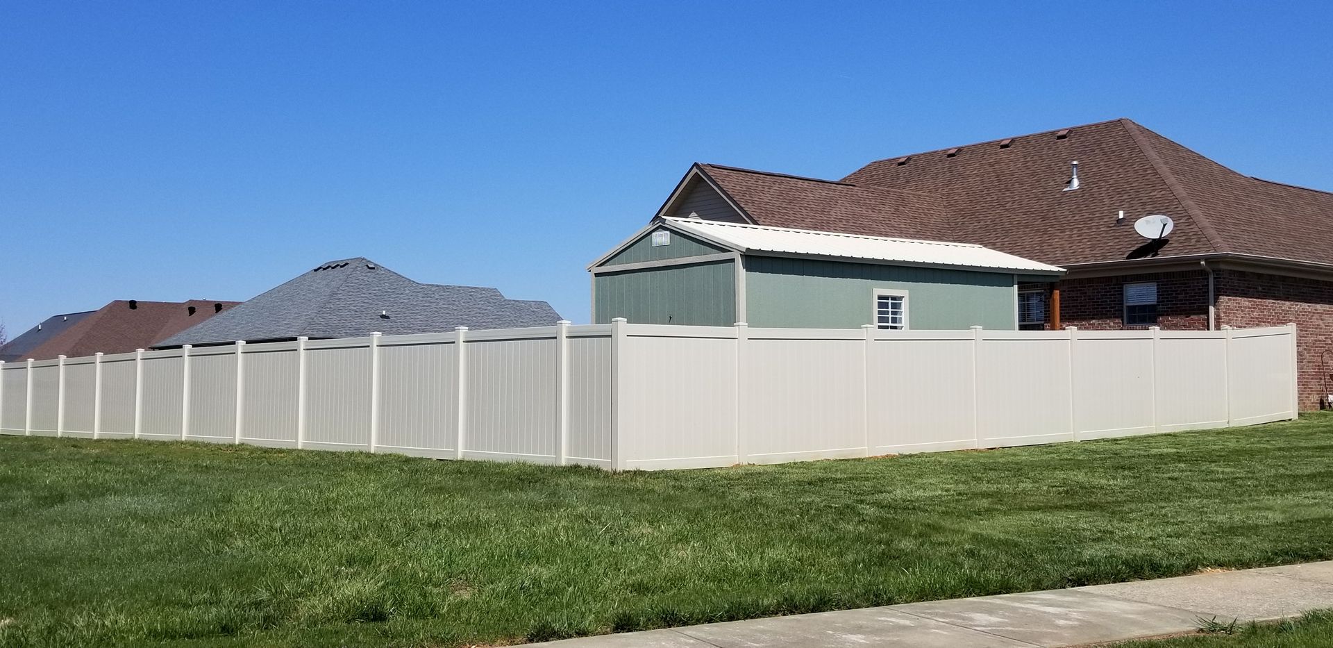 White fence surrounds green lawn with houses in the background on a sunny day.