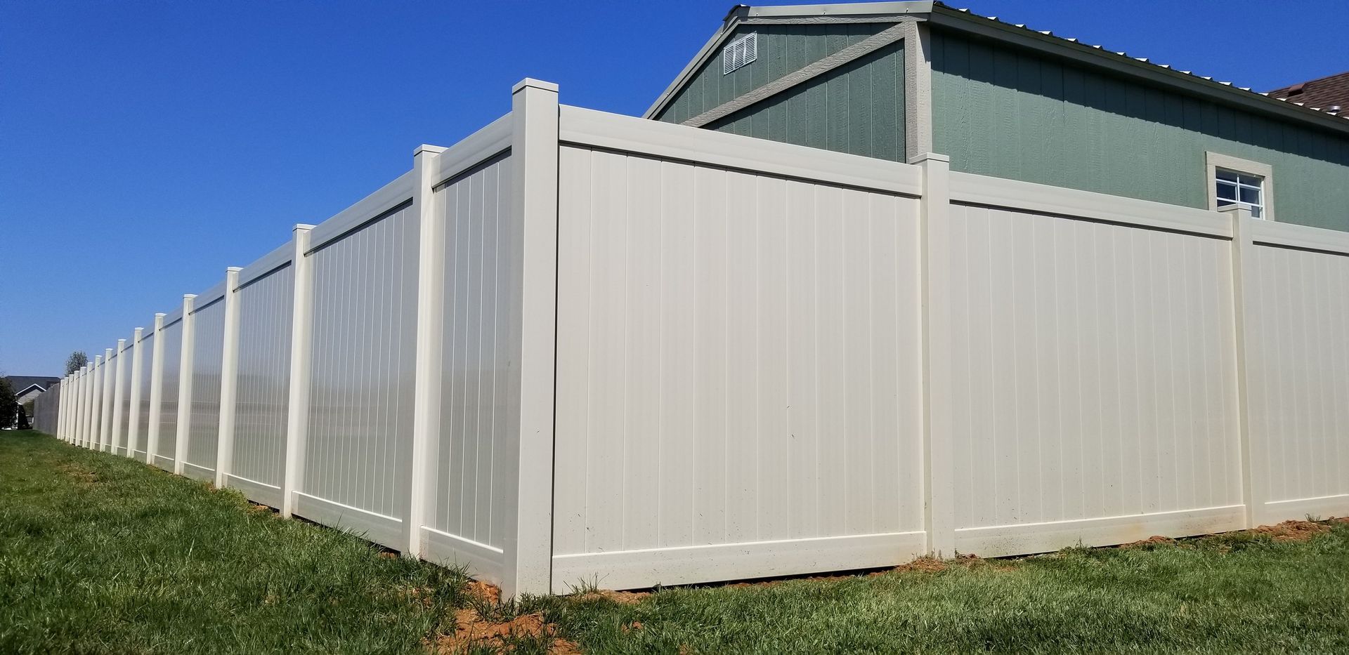 Beige vinyl fence around a grassy yard, with a blue sky and part of a green house in the background.