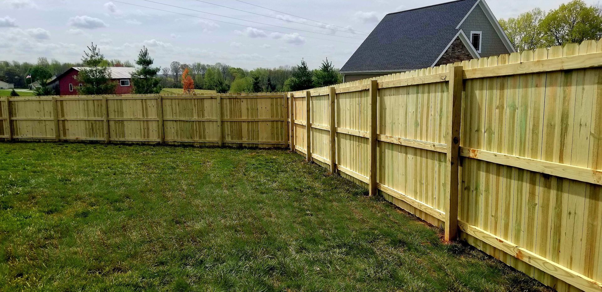 A wooden fence surrounds a grassy yard, with a house in the background on a sunny day.