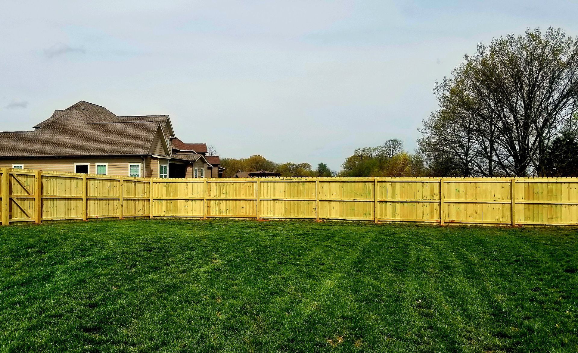Wooden fence bordering a grassy yard with a house and trees in the background under a cloudy sky.