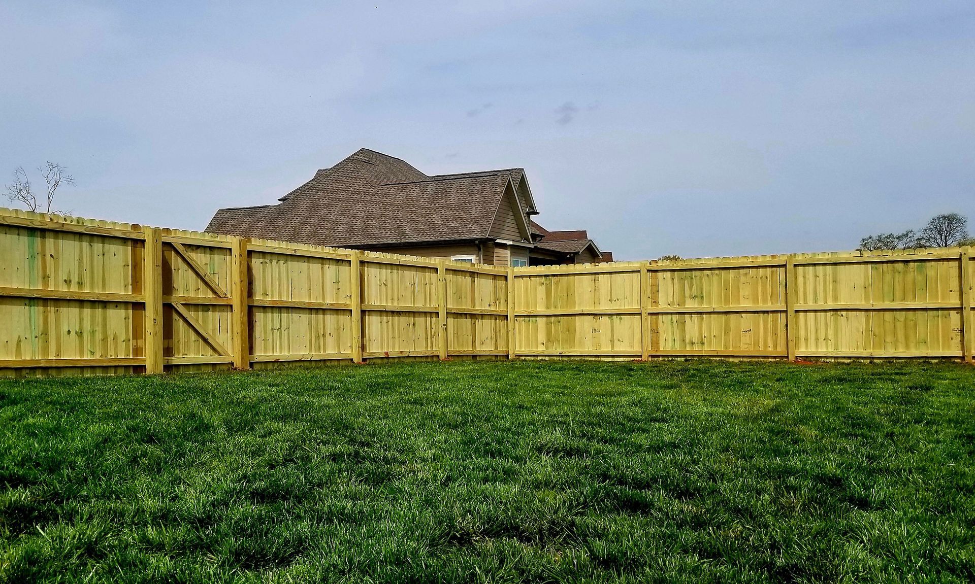 Wooden fence surrounding a grassy yard with a house visible in the background under a cloudy sky.