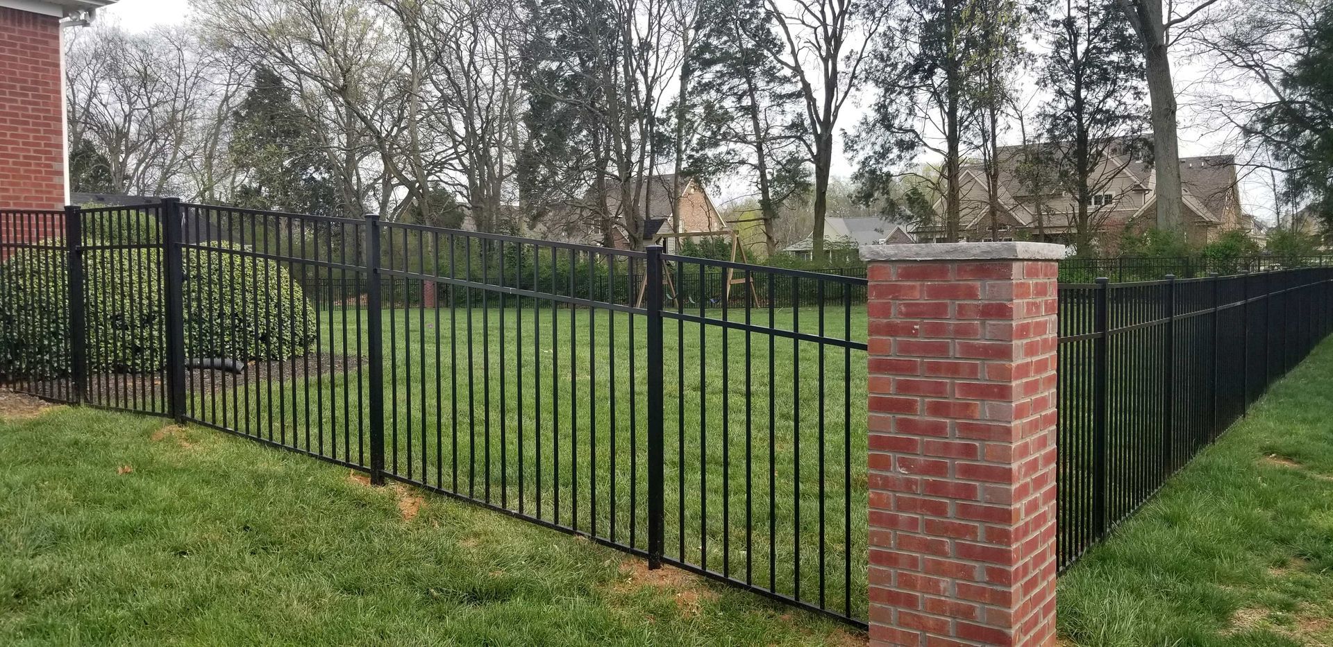 Black metal fence with brick pillars on a grassy lawn, trees in the background.