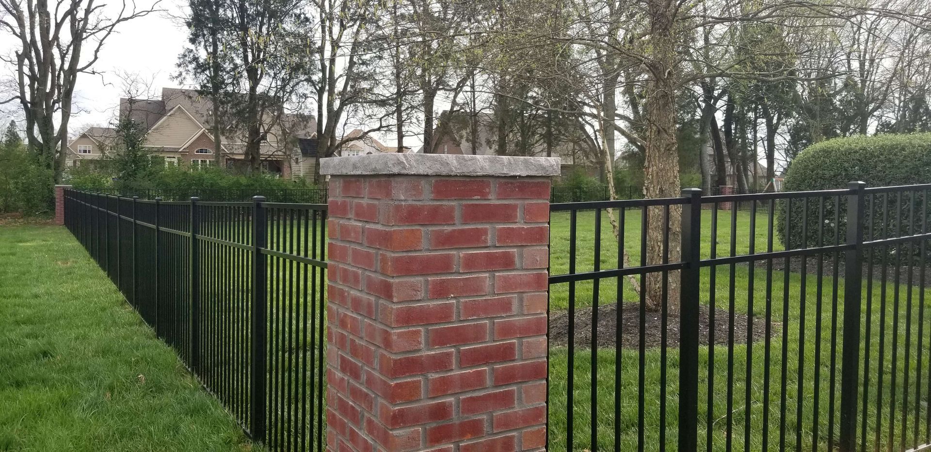 Black metal fence with brick pillar, green grass, trees, and a house in the background.