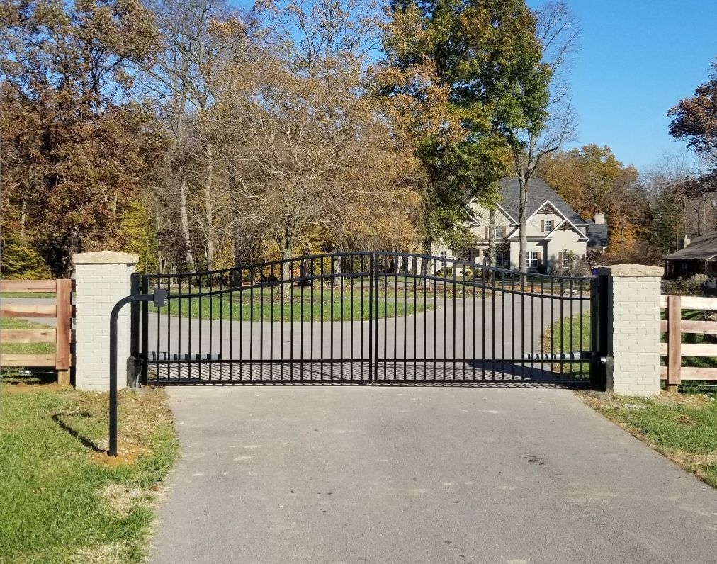 Black metal gate across a paved driveway, flanked by stone columns and wooden fences, leading to a house.