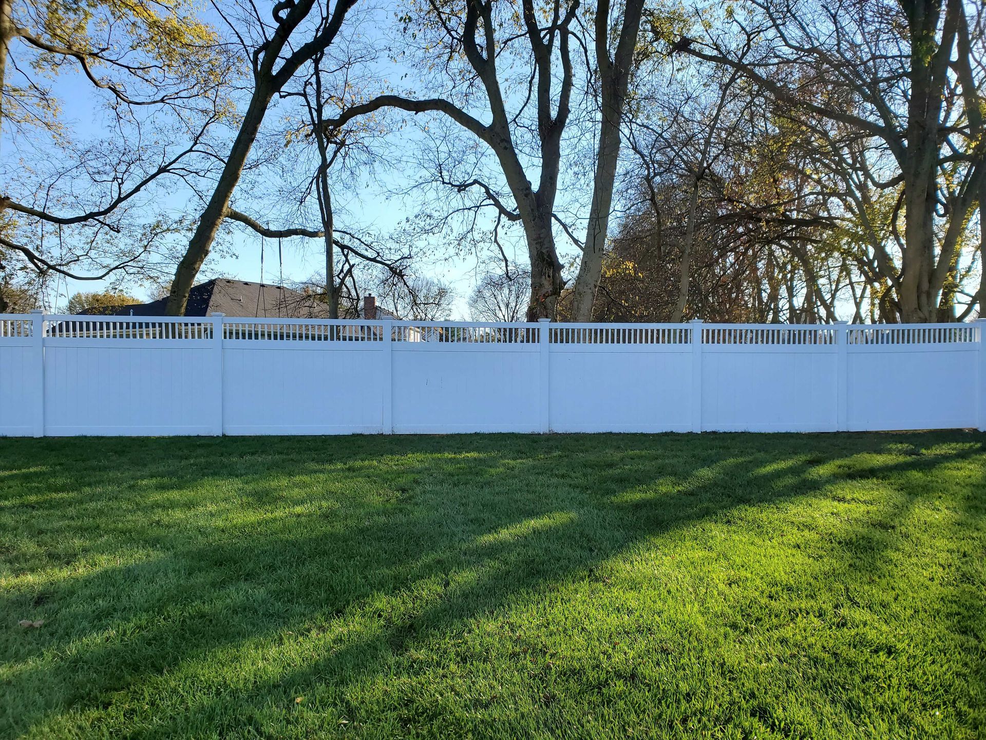White fence bordering a green lawn, with trees and a blue sky in the background.