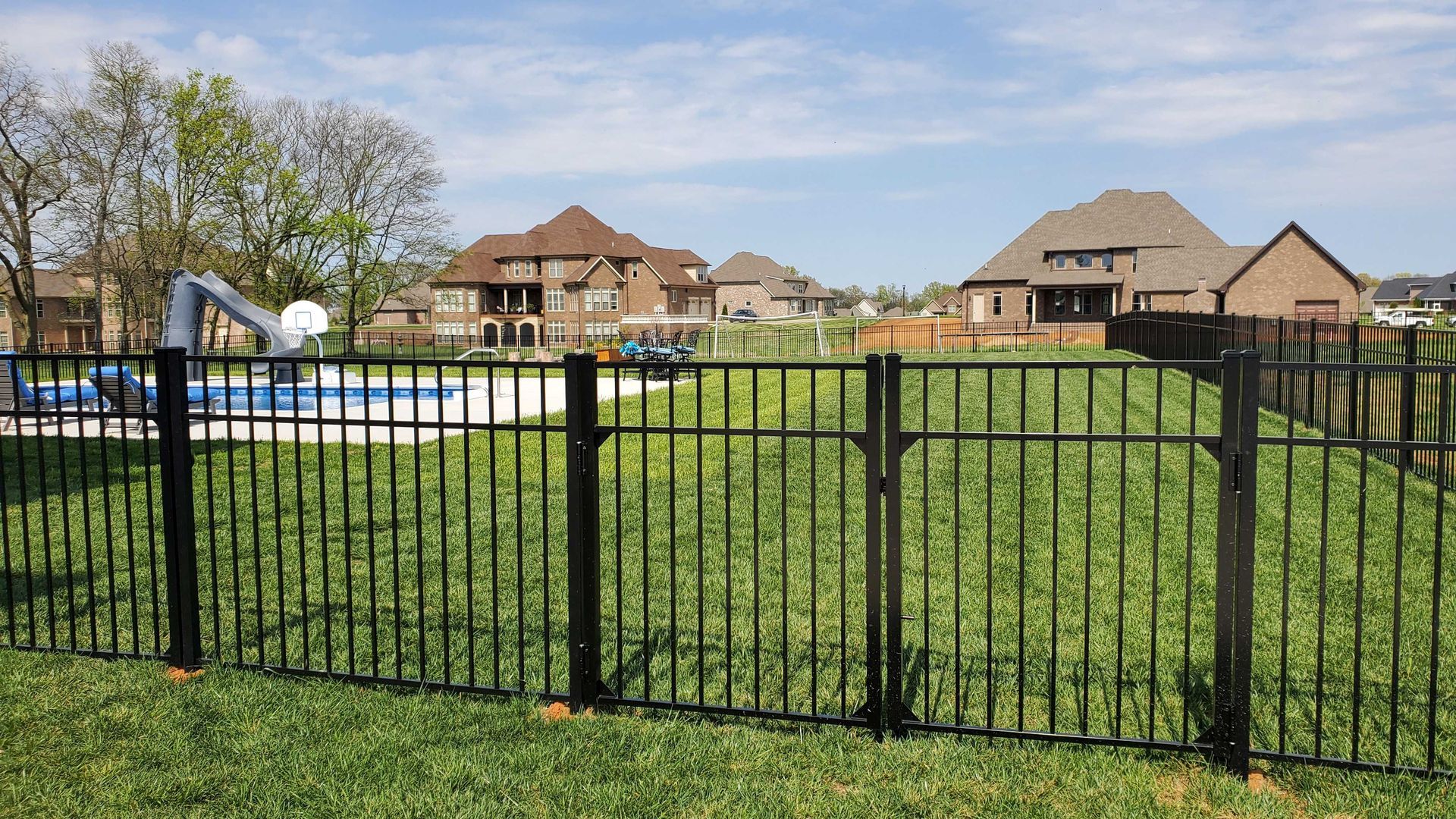 Black metal fence surrounding a grassy backyard with a pool, houses in background under blue sky.