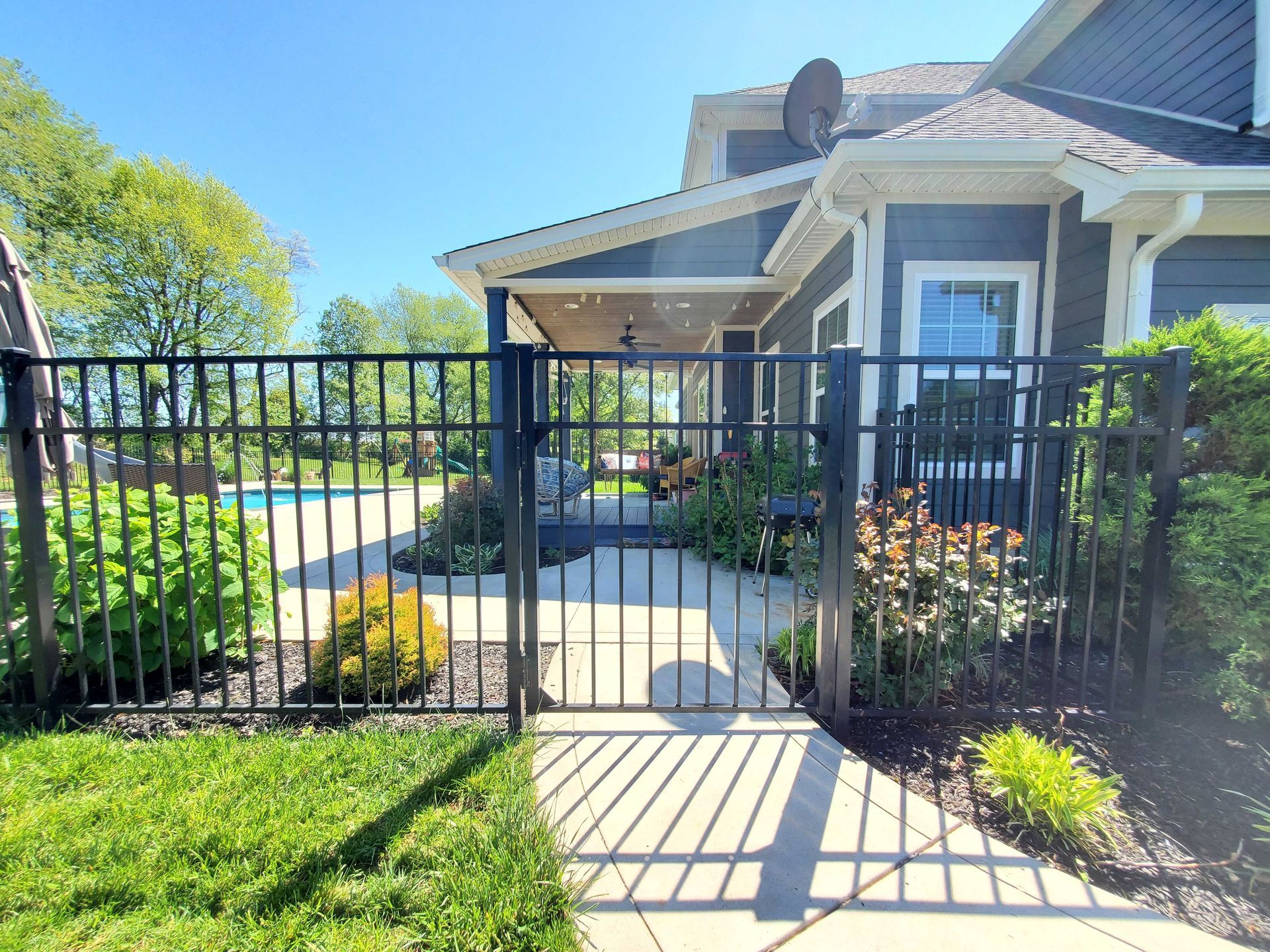 Black metal fence with gate in front of a house with a pool visible in the distance. Sunny day.