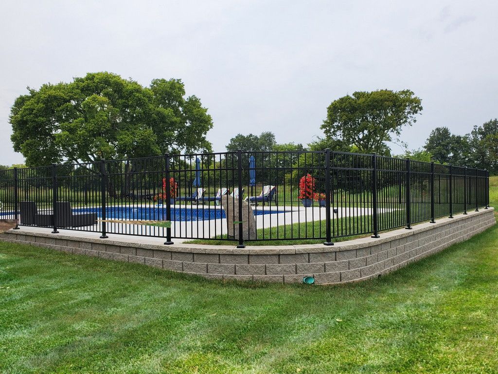 Black fence encloses a pool area on a stone retaining wall, with trees and a cloudy sky in the background.