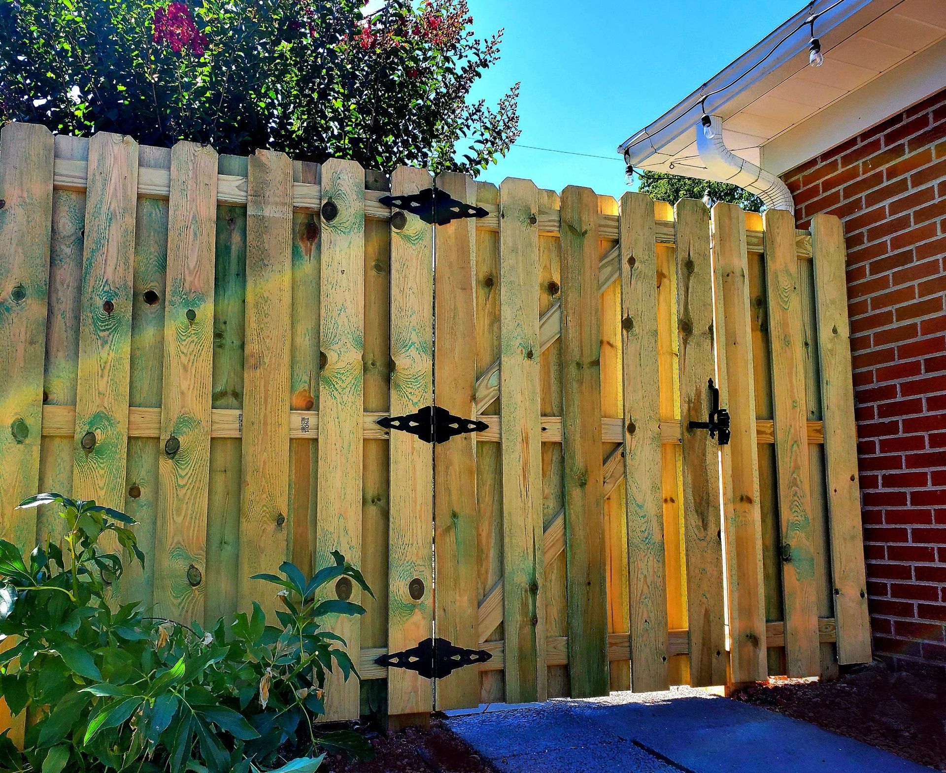 Wooden picket fence with gate, black hinges and latch, against a brick building, sunny outdoors.