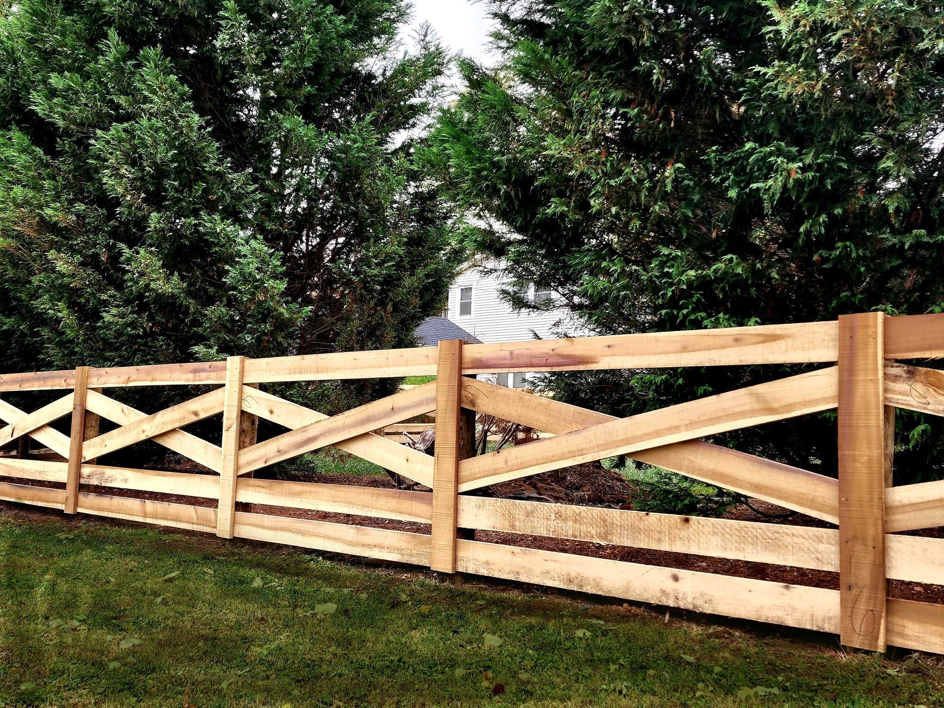 Wooden fence with crisscross design, bordering a green lawn with trees and a house in the background.