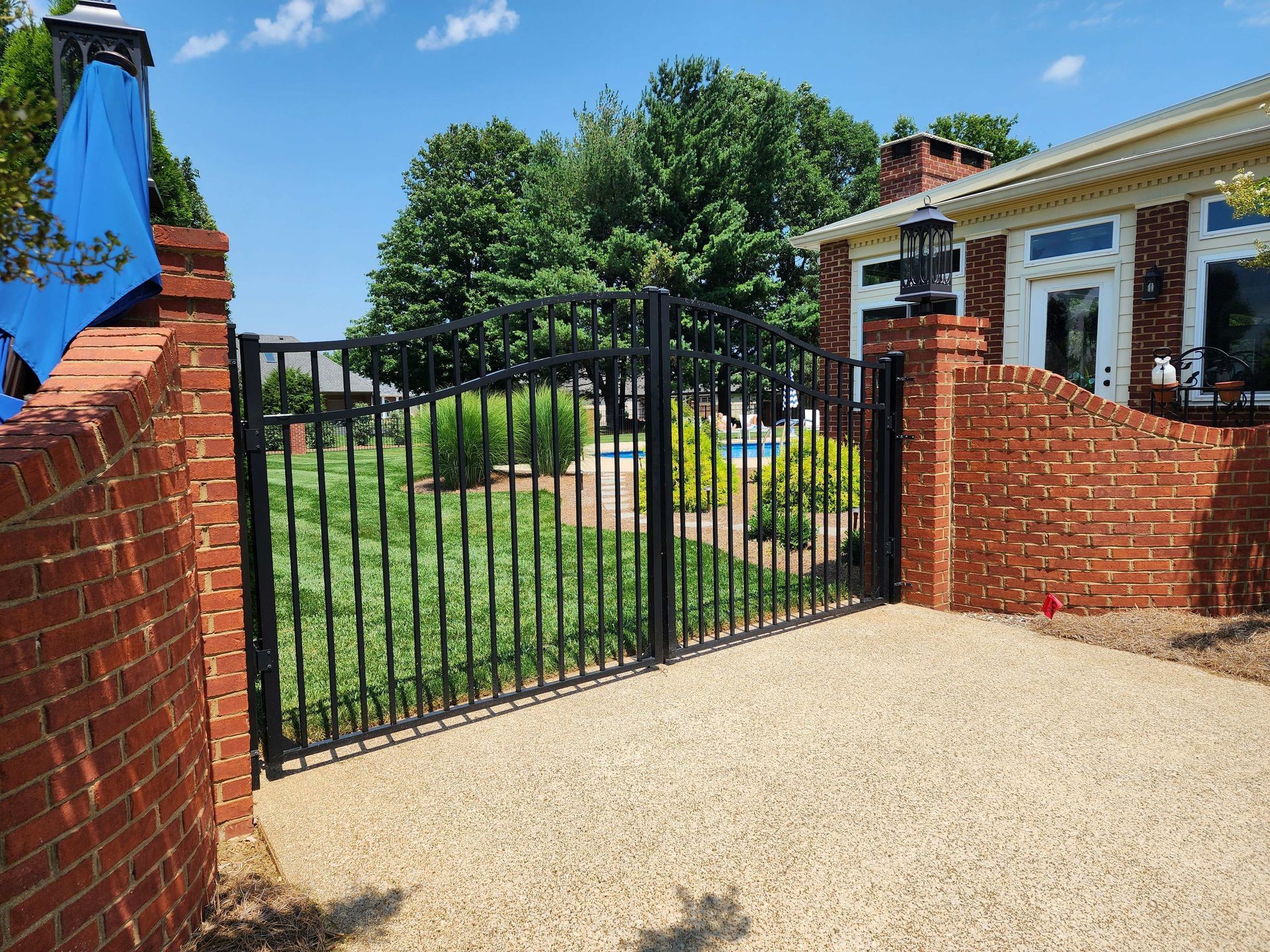 Black iron gate between brick walls, leading to a backyard with green grass and trees.