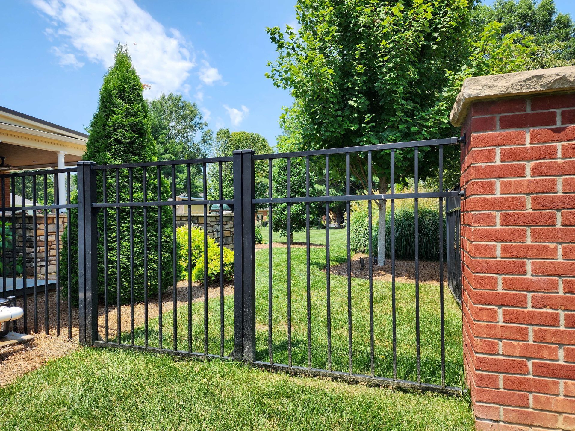 Black metal fence section next to a red brick column, with a grassy yard and trees in the background.