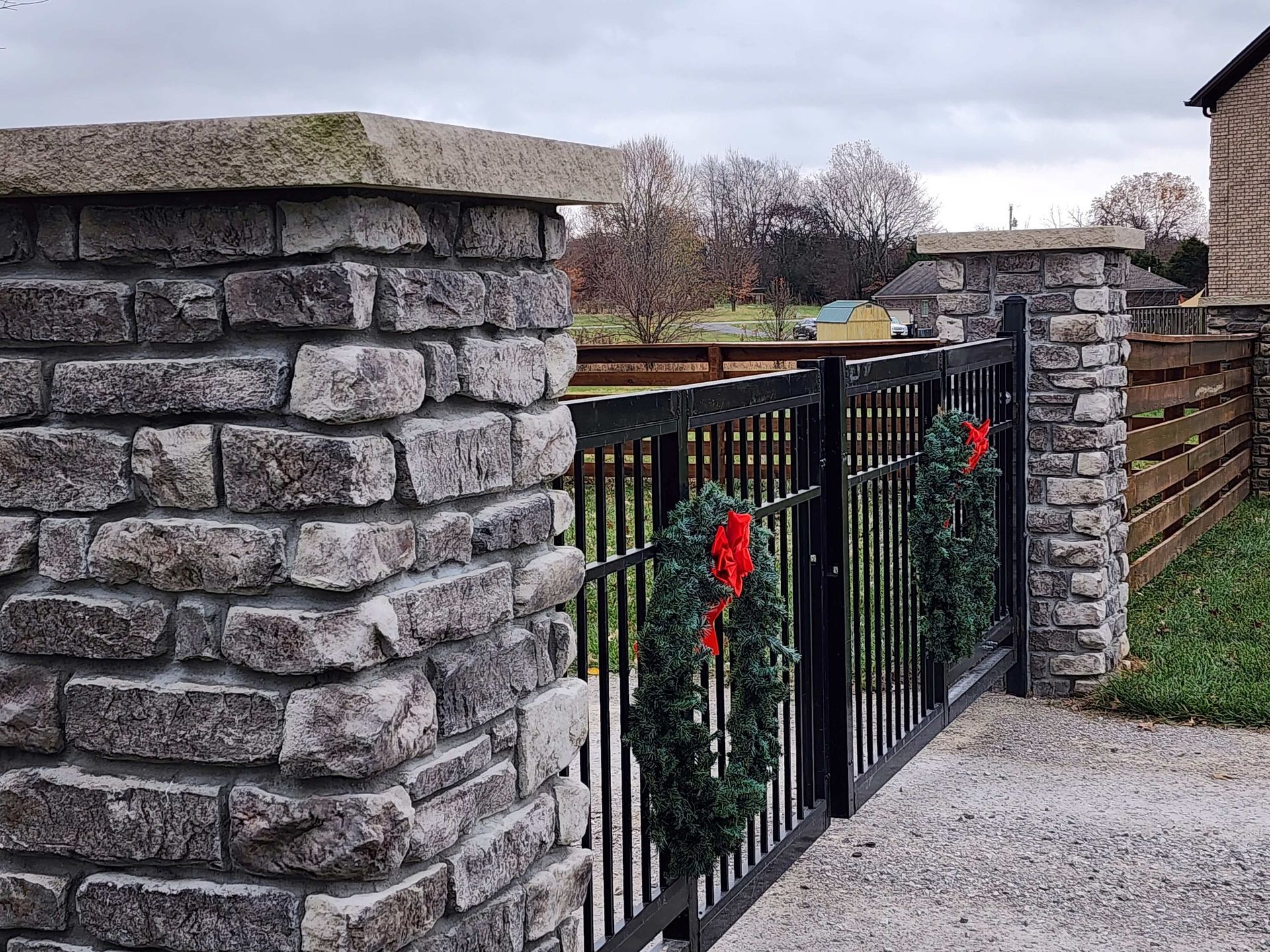 Stone pillars flank a black metal gate adorned with wreaths. A wooden fence and field are in the background.