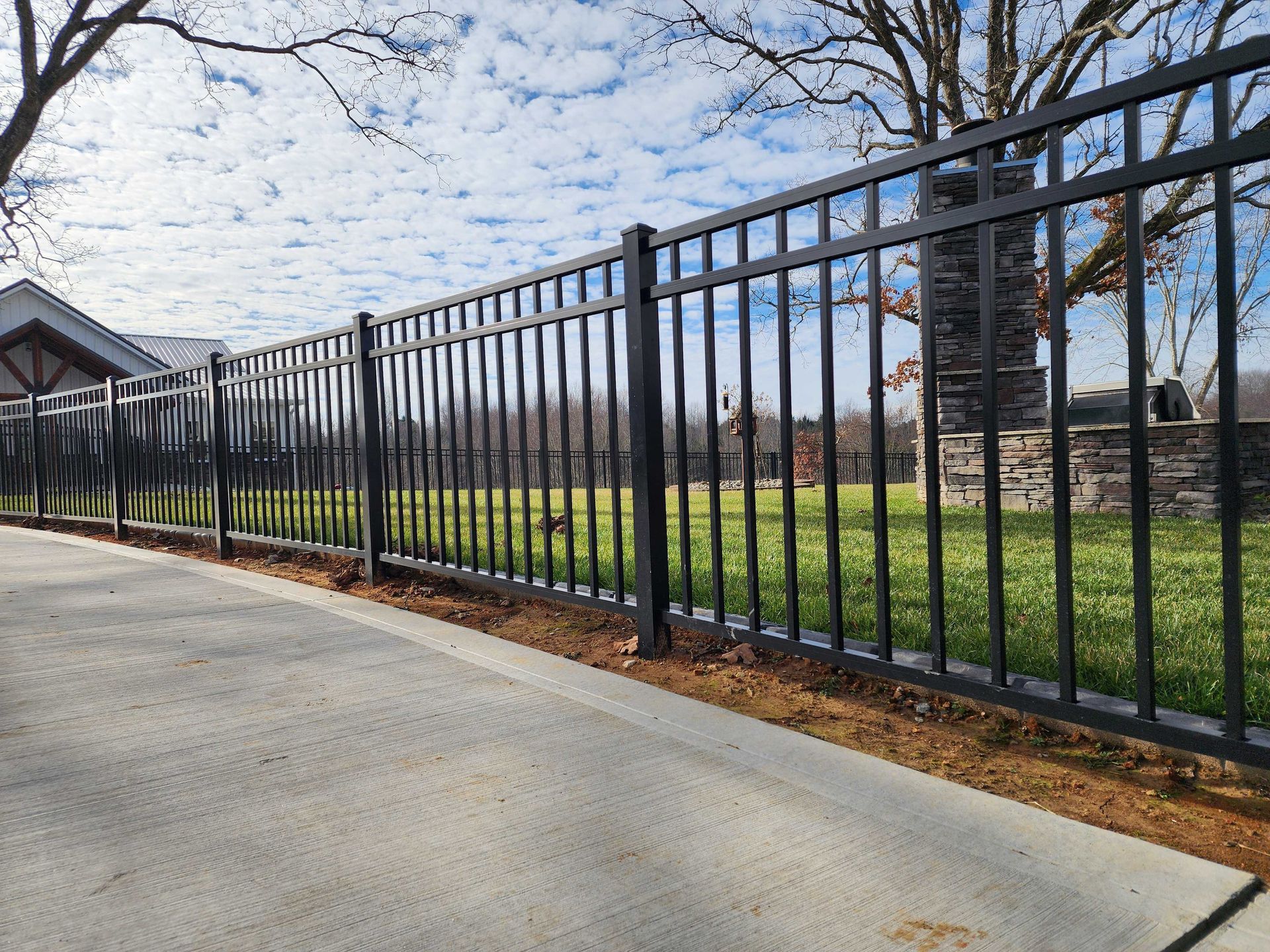 Black metal fence along a concrete path and grassy lawn under a partly cloudy sky.