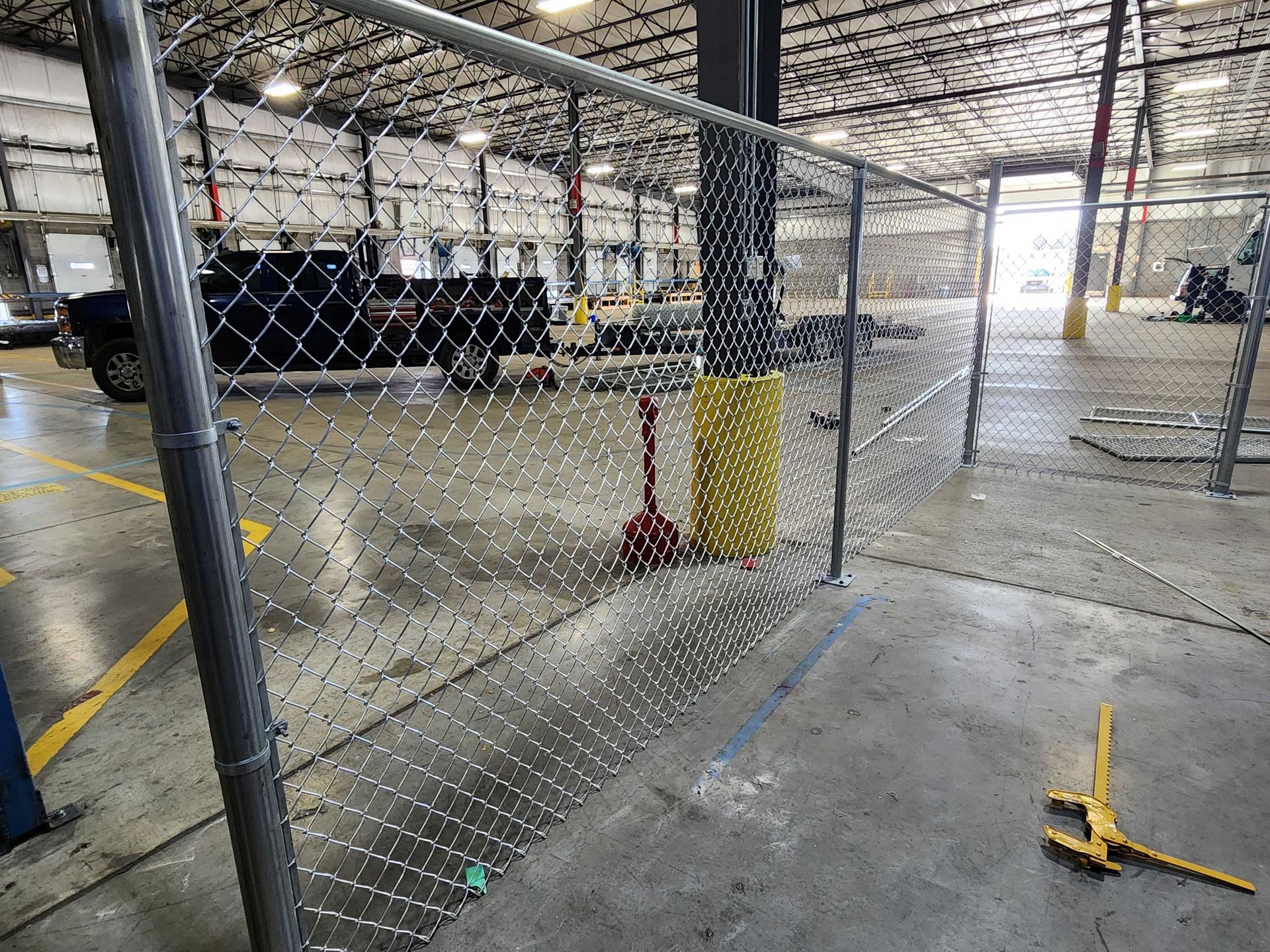 Chain-link fence inside a warehouse, separating a work area. A vehicle and equipment are visible in the background.