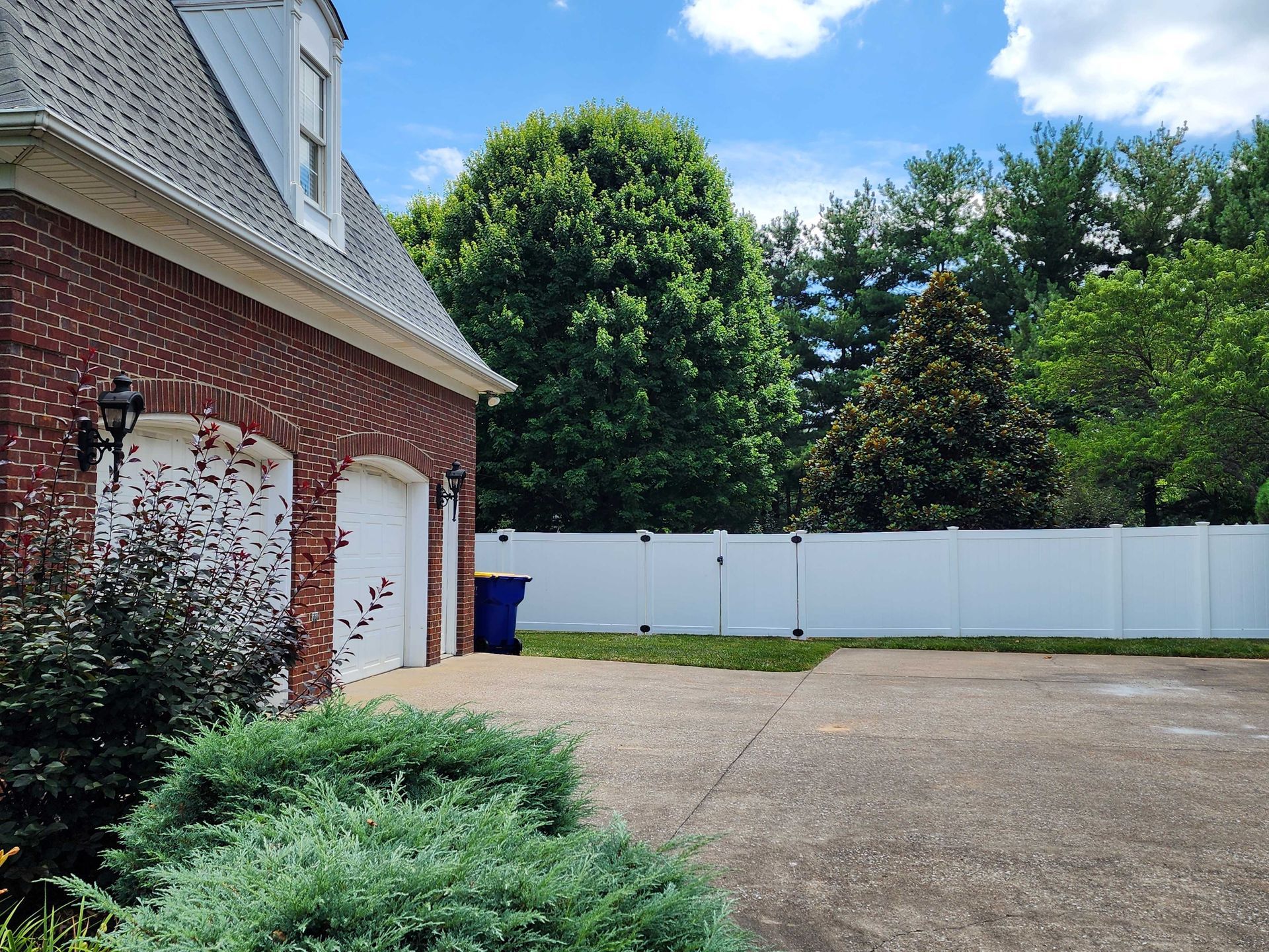 Brick garage with white doors, blue trash can, and white fence against trees.